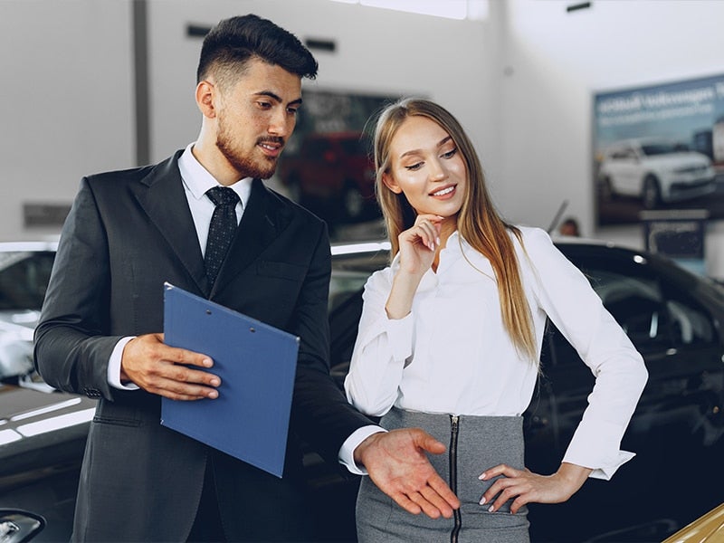 A car salesman with a clipboard shows a vehicle to a woman