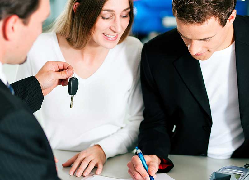 A man signs paperwork while a car salesman holds up vehicle keys
